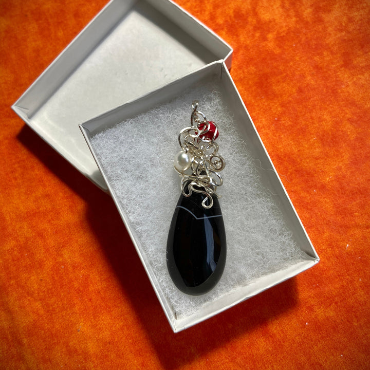 Black teardrop Agate pendant, silver-plated wire wrap and red gemstone in a box on an orange surface