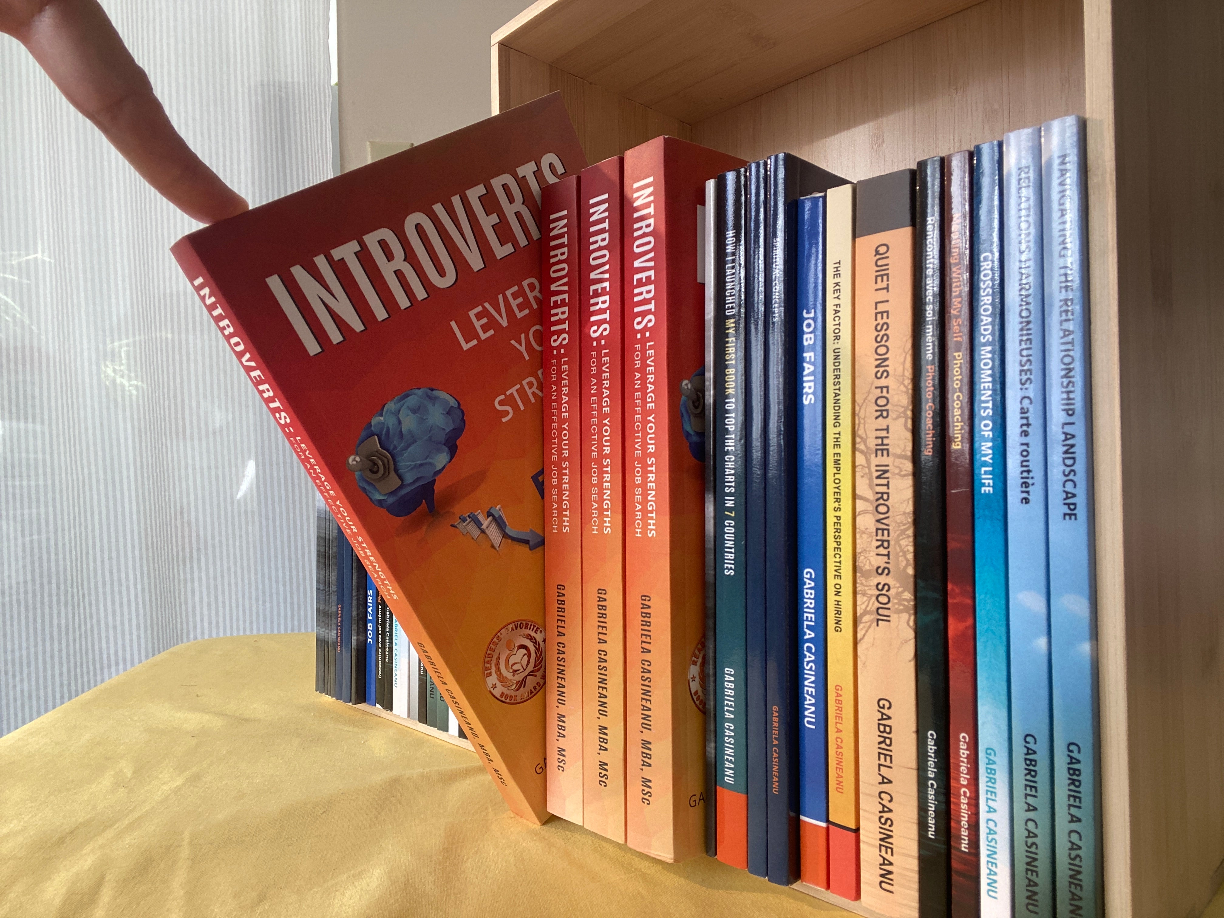 Bookshelf with books by Gabriela Casineanu, including an orange one titled 'Introverts' held by a hand.