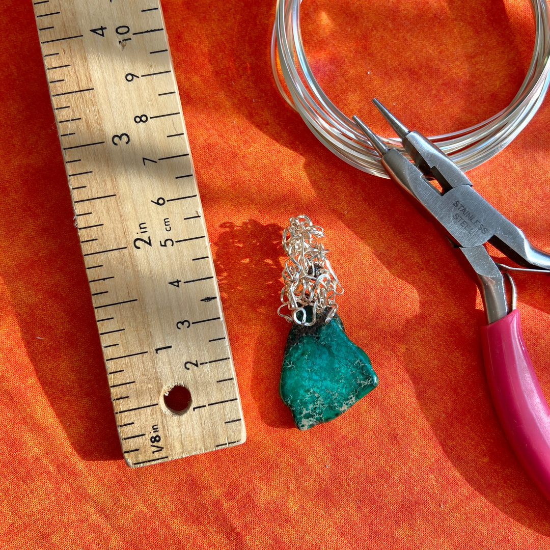 Jewelry-making setup with a blue-green stone pendant, silver wire, pliers, and ruler on an orange surface.