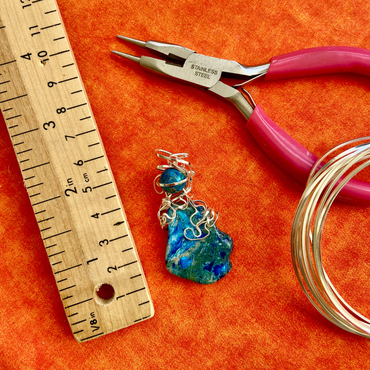 Jewelry-making setup with a blue pendant, pliers, and wire on an orange surface.