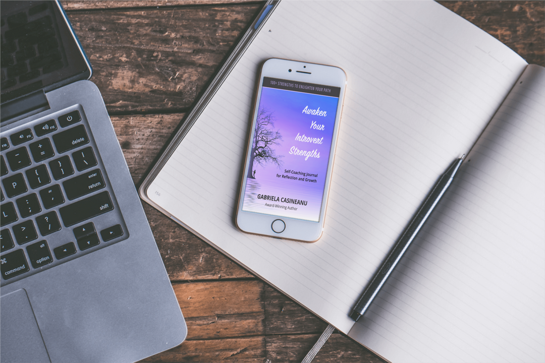 Phone with Awaken your Introvert Strengths ebook cover displayed on a wooden desk with a laptop and notebook.