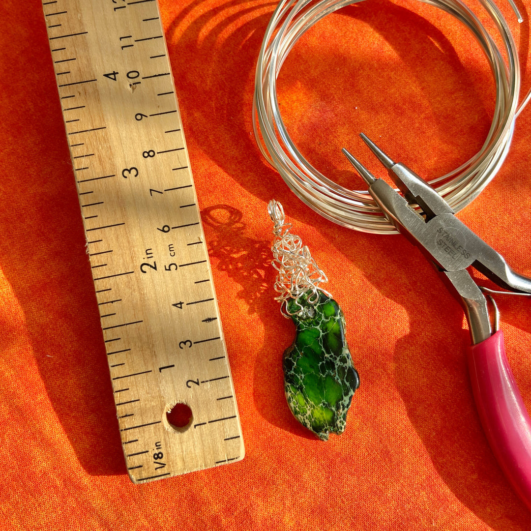 Green gemstone pendant with silver wire and pliers on an orange surface, next to a wooden ruler.