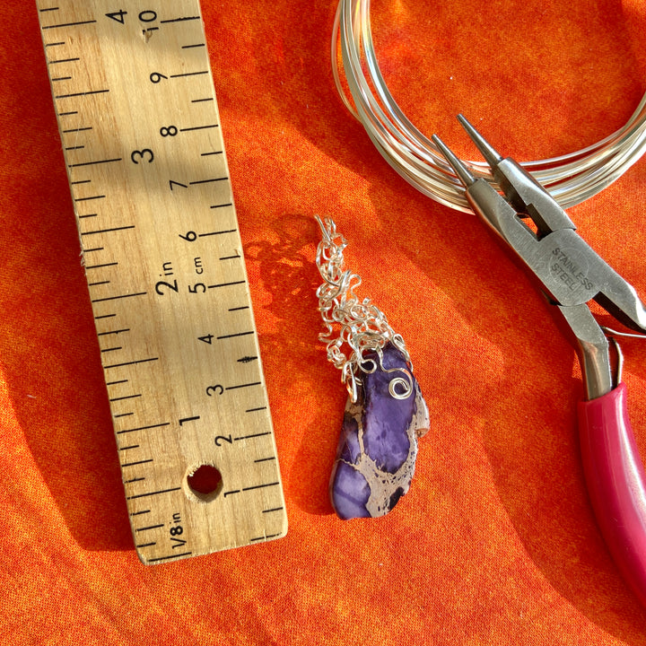 Jewelry pendant with purple stone and silver chain on an orange surface with a ruler and pliers.