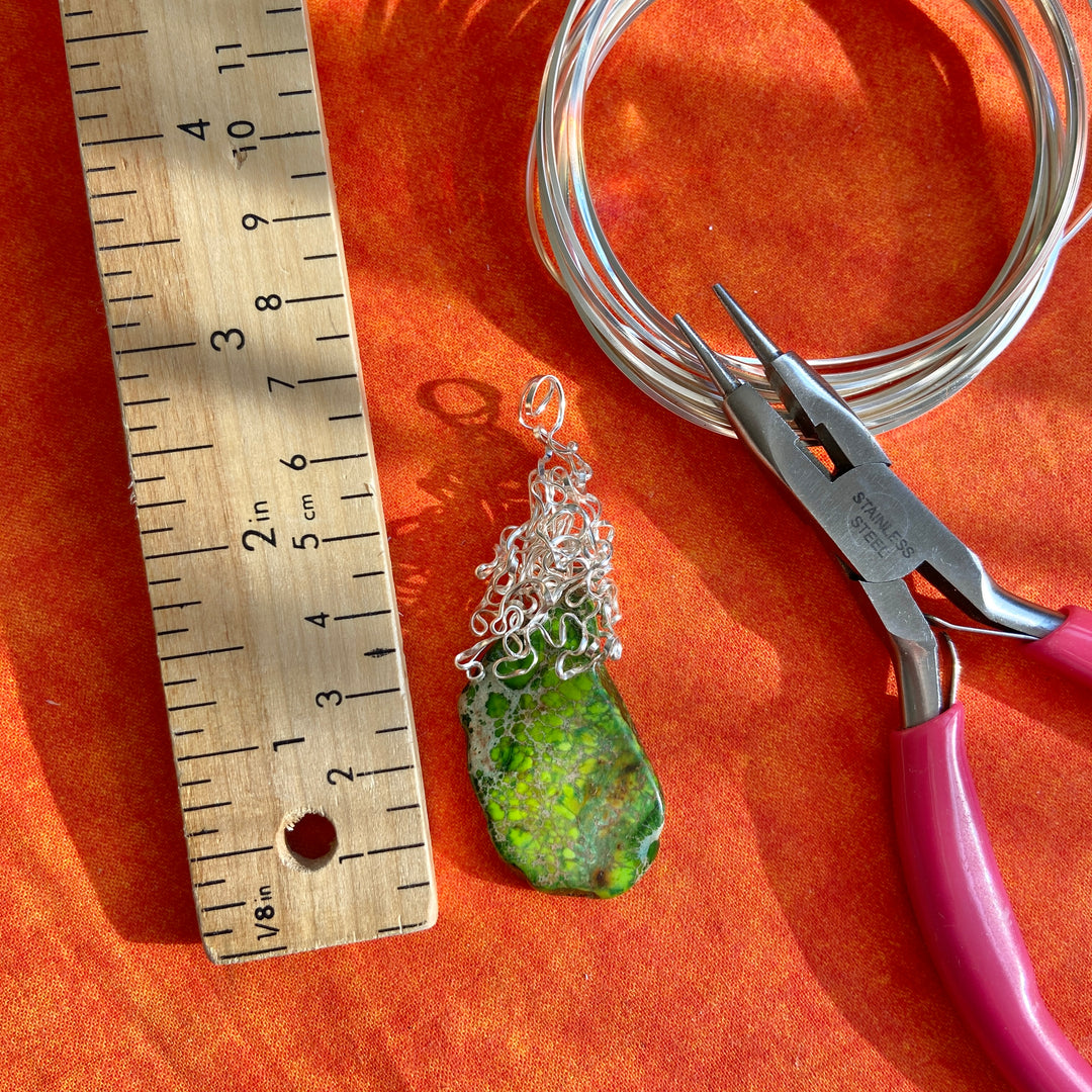 Green pendant with silver chain on an orange surface with a ruler and pliers.