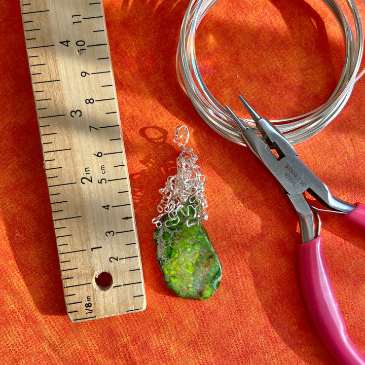 Green pendant with silver chain on an orange surface with a ruler and pliers.