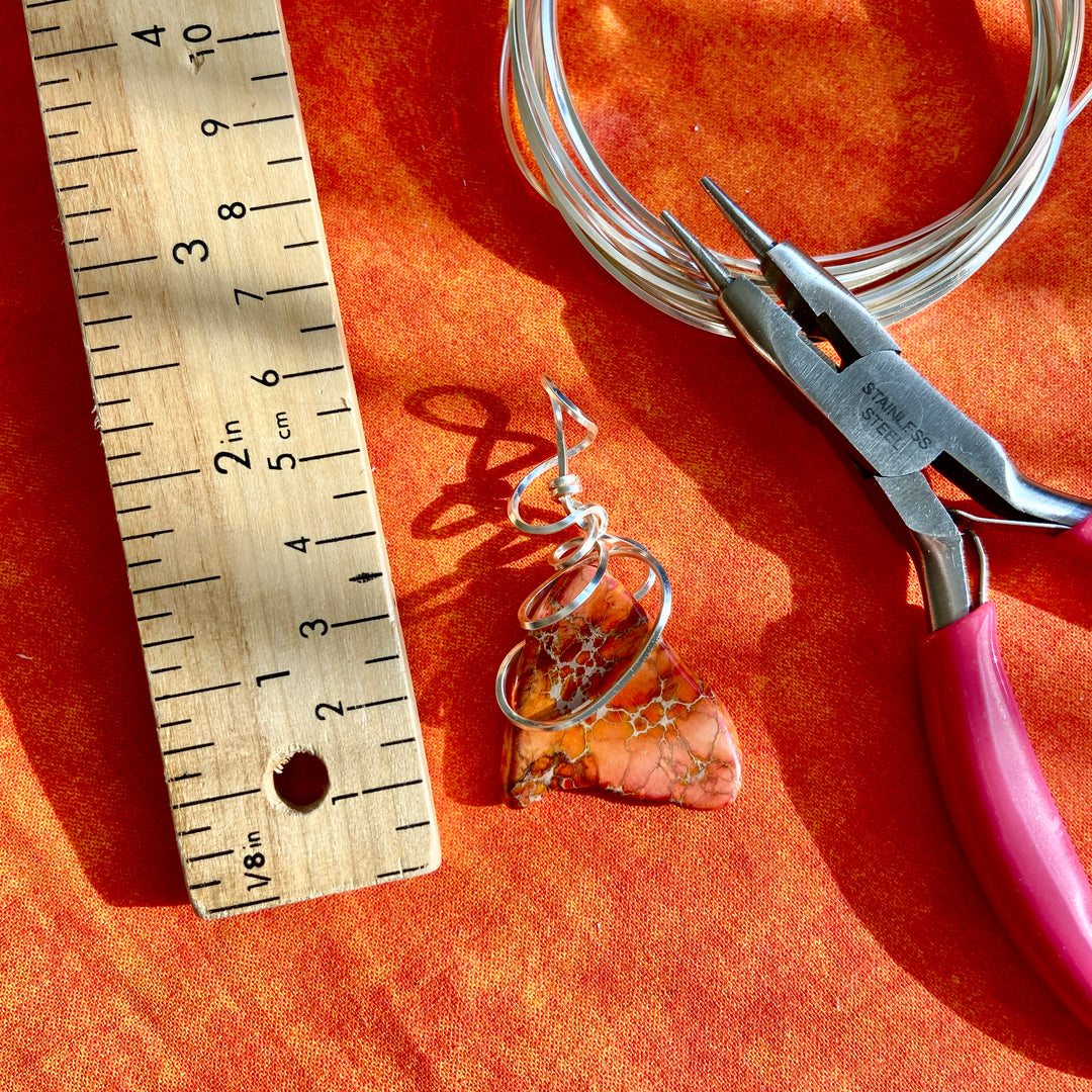 Handmade pendant with wire wrapping technique on an orange fabric background, next to a ruler and pliers.