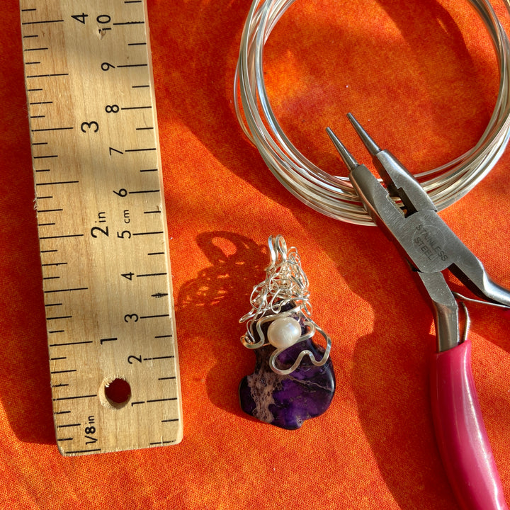 Jewelry-making tools and materials including wire, pliers, and a purple pendant on an orange surface with a ruler for scale.