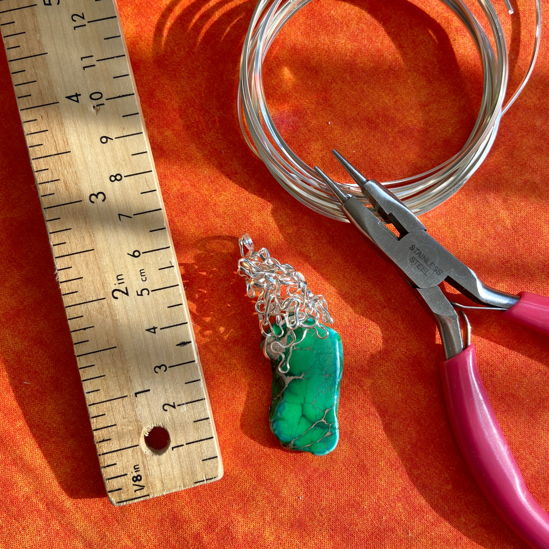 Jewelry-making tools and materials including a turquoise pendant, silver chain, pliers, and a ruler on an orange surface.