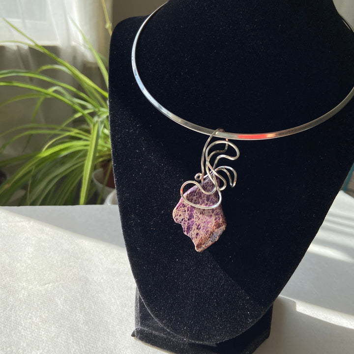 Necklace with a purple stone pendant on a black stand with a plant in the background